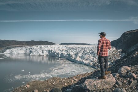 A young man traveler tourist standing in front of Eqip Sermia glacier called Eqi Glacier. Wall of ice in background. The concept of global warming and professional guides. On a sunny day with blue sky.の写真素材