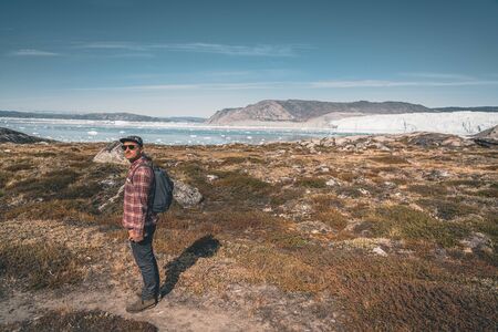 A young man traveler tourist standing in front of Eqip Sermia glacier called Eqi Glacier. Wall of ice in background. The concept of global warming and professional guides. On a sunny day with blue sky.の写真素材
