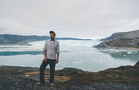 Happy and smiling young man standing in front of Eqip Sermia glacier called Eqi Glacier. Tourist with huge wall of ice in background. The concept of global warming and professional guides. Overcast day.の写真素材