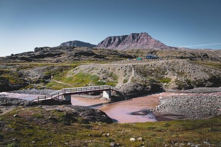 Hiking path on arctic Disko island in Greenland. River bed with bridge and colourful houses in background. Table mountains on a sunny day in summer.のeditorial素材