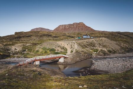 Hiking path on arctic Disko island in Greenland. River bed with bridge and colourful houses in background. Table mountains on a sunny day in summer.のeditorial素材