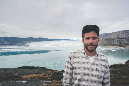 Happy and smiling young man standing in front of Eqip Sermia glacier called Eqi Glacier. Tourist with huge wall of ice in background. The concept of global warming and professional guides. Overcast day.の写真素材