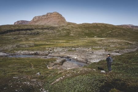 Young man hiker in the arctic landscape of Disko Bay in Greeland in Summer. Blue Sky and green meadows. Arctic Circle Trail with temple mountains. Disko Island and Village of Qeqertarsuaq, Photo taken in Greenland.の写真素材