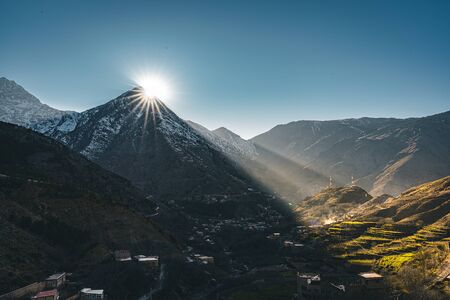 A view of sunset sunrise with sunstar in rural mountain village Imlil in High Atlas mountains Morocco in Africa. Snow covered peaks in background with lightrays and blue sky.の写真素材