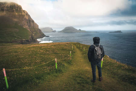 Young man tourist standing on Faroe islands with stormy view towards mykines on the island of Vagar as seen from mulafossur waterfall. Low clouds with atlantic ocean waves.の写真素材