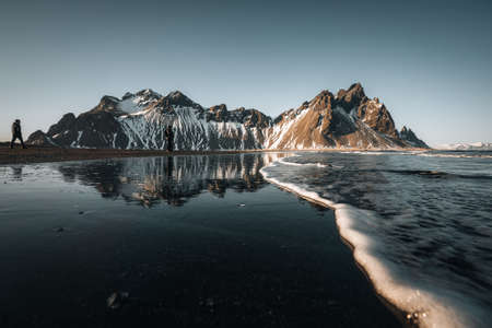 Young man tourist standing at beach during sunset runrise and gorgeous reflection of Vestrahorn mountain on Stokksnes cape in Iceland. Beautiful snow covered mountains. Location: Stokksnes cape, Vestrahorn Batman Mount , Iceland, Europe. Photo taken in Iceland.の写真素材
