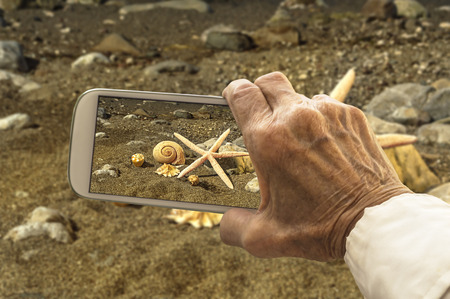 Old hand takes a picture of starfish and sea shells on smart phone. Selective focus.の写真素材