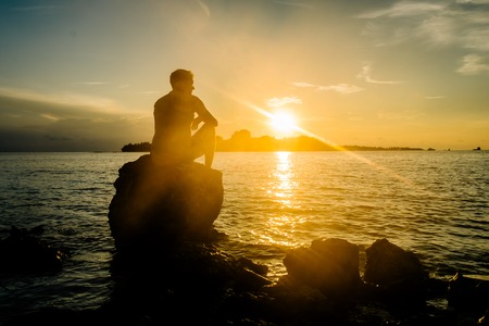 Handsome man sitting on a rock on the beach and watching the sunset, Maldivesの写真素材