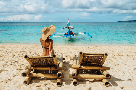 Woman with blue hat relaxing on the bamboo chair on a beautiful white beach. Philippines. Boat on the backgroundの写真素材