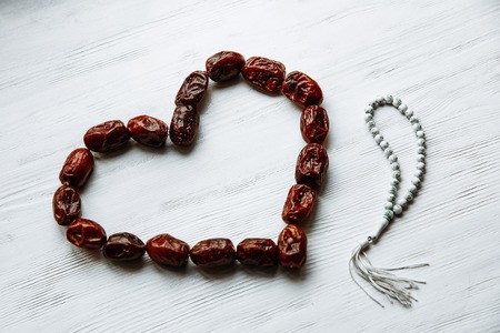 heart shaped by dates and white rosary on the white wooden table for Ramadan and Friday message...