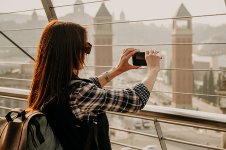 Young girl with long brown hair standing on view point and taking a picture of the city landscape using mobile phone. Backside view. Monjuic, Barcelona, Spainの写真素材