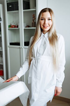 portrait of smiling cosmetologist in white coat standing at empty massage table in salonの写真素材