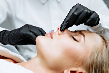 The doctor cosmetologist makes prick in the nose to correct the hump of a beautiful woman in a beauty salon. Cosmetology skin care.の写真素材