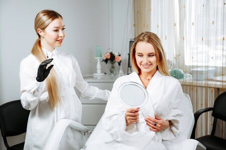 Young female client received cosmetic face treatments in clinic of aesthetic medicine, looking in mirror at resultsの写真素材
