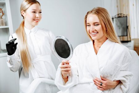 Young female client received cosmetic face treatments in clinic of aesthetic medicine, looking in mirror at resultsの写真素材
