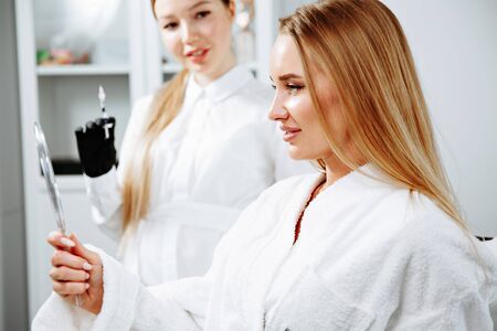 Young female client received cosmetic face treatments in clinic of aesthetic medicine, looking in mirror at resultsの写真素材