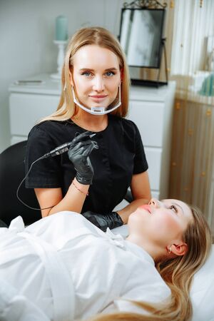 Master beautician, blonde, bent over lying European girl and makes her permanent lip makeup in the beauty salon. Close up. The view from the top. Vertical orientation of the imageの写真素材