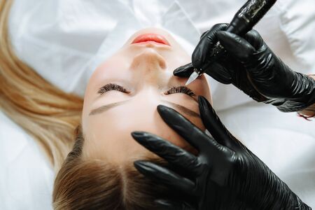 Close up of woman lying on couch at beauty salon with closed eyes and enjoying when cosmetologist making eyeliner permanent make up. Professional wearing black gloves and holding speciality tool.の写真素材