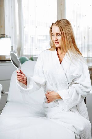 Beautiful young woman is looking into the mirror and smiling while sitting at the cosmeticianの写真素材