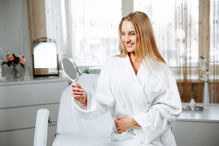 Beautiful young woman is looking into the mirror and smiling while sitting at the cosmeticianの写真素材