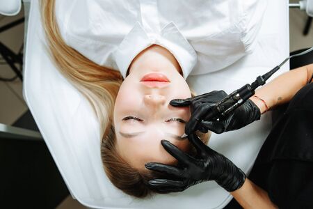 Close up of woman lying on couch at beauty salon with closed eyes and enjoying when cosmetologist making eyeliner permanent make up. Professional wearing black gloves and holding speciality tool.の写真素材