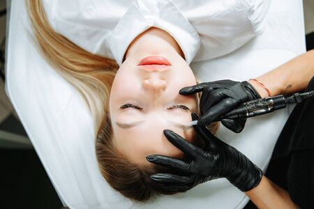 Close up of woman lying on couch at beauty salon with closed eyes and enjoying when cosmetologist making eyeliner permanent make up. Professional wearing black gloves and holding speciality tool.の写真素材