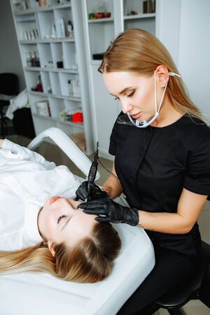 Master beautician, blonde, bent over lying European girl and makes her permanent lip makeup in the beauty salon. Close up. The view from the top. Vertical orientation of the imageの写真素材