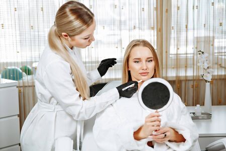 Young female client received cosmetic face treatments in clinic of aesthetic medicine, looking in mirror at resultsの写真素材