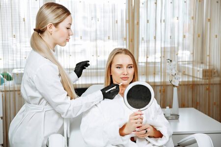 Young female client received cosmetic face treatments in clinic of aesthetic medicine, looking in mirror at resultsの写真素材