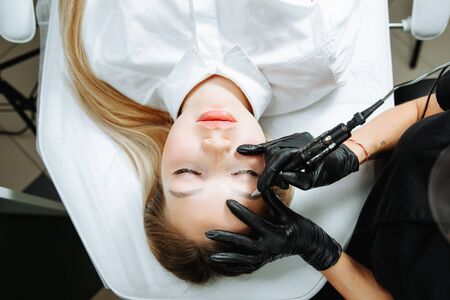 Close up of woman lying on couch at beauty salon with closed eyes and enjoying when cosmetologist making eyeliner permanent make up. Professional wearing black gloves and holding speciality tool.の写真素材