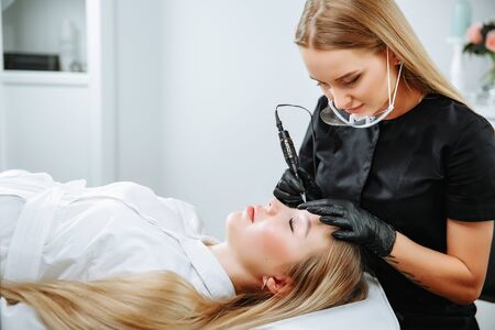 Master beautician, blonde, bent over lying European girl and makes her permanent lip makeup in the beauty salon. Close up. The view from the top. Vertical orientation of the imageの写真素材