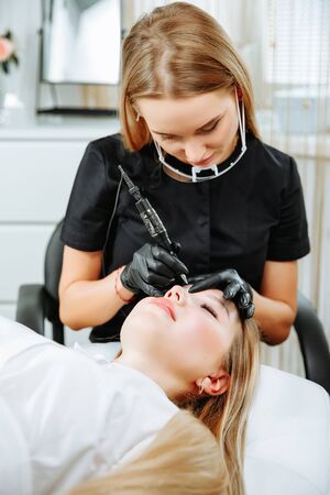 Master beautician, blonde, bent over lying European girl and makes her permanent lip makeup in the beauty salon. Close up. The view from the top. Vertical orientation of the imageの写真素材