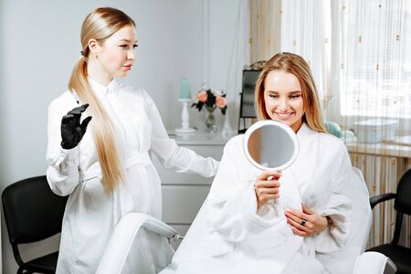 Young female client received cosmetic face treatments in clinic of aesthetic medicine, looking in mirror at resultsの写真素材