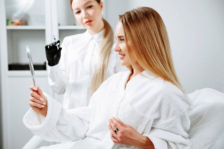 Young female client received cosmetic face treatments in clinic of aesthetic medicine, looking in mirror at resultsの写真素材