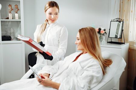 Consultation in cosmetology clinic. Female beauty doctor talking with patient. A young girl tells what she wants change in her face. Client shows on himself. Doctor listening carefullyの写真素材