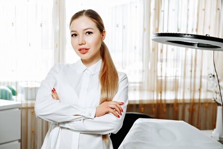 portrait of smiling cosmetologist in white coat standing at empty massage table in salonの写真素材
