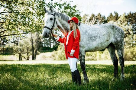 beauty blondie woman in red jacket and red hat with horse in the field, effect of toning. Equestrian sport concept. Fashion photoshootの写真素材