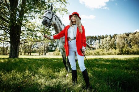 beauty blondie woman in red jacket and red hat with horse in the field, effect of toning. Equestrian sport concept. Fashion photoshootの写真素材