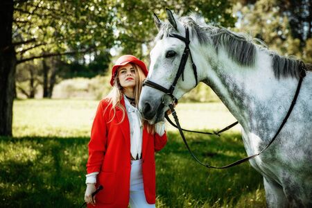beauty blondie woman in red jacket and red hat with horse in the field, effect of toning. Equestrian sport concept. Fashion photoshootの写真素材