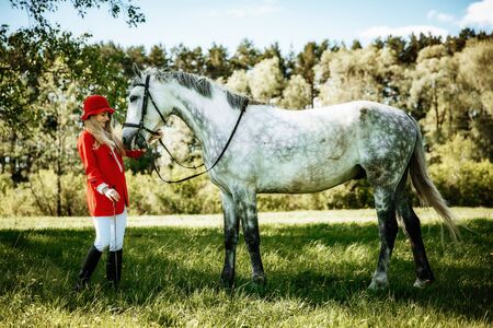 beauty blondie woman in red jacket and red hat with horse in the field, effect of toning. Equestrian sport concept. Fashion photoshootの写真素材