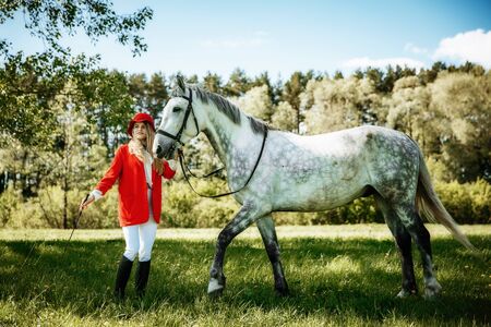 beauty blondie woman in red jacket and red hat with horse in the field, effect of toning. Equestrian sport concept. Fashion photoshootの写真素材