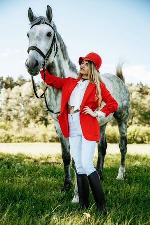 Beautiful young girl with blond hair in a red jacket with a fringe , wearing red hat, smiling and stroking her horse in summer field. equestrian sport conceptの写真素材