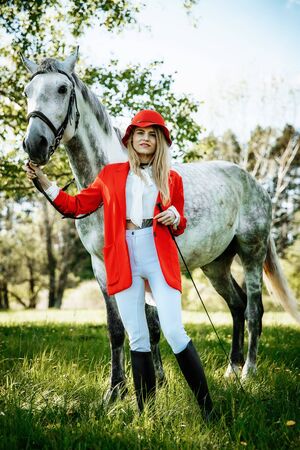beauty blondie woman in red jacket and red hat with horse in the field, effect of toning. Equestrian sport concept. Fashion photoshootの写真素材