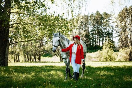 Beautiful young girl with blond hair in a red jacket with a fringe , wearing red hat, smiling and stroking her horse in summer field. equestrian sport conceptの写真素材