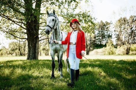 Beautiful young girl with blond hair in a red jacket with a fringe , wearing red hat, smiling and stroking her horse in summer field. equestrian sport conceptの写真素材