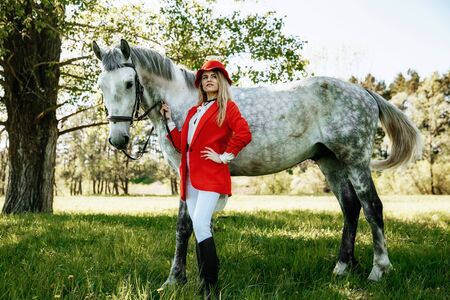 Beautiful young girl with blond hair in a red jacket with a fringe , wearing red hat, smiling and stroking her horse in summer field. equestrian sport conceptの写真素材