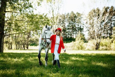 Beautiful young girl with blond hair in a red jacket with a fringe , wearing red hat, smiling and stroking her horse in summer field. equestrian sport conceptの写真素材
