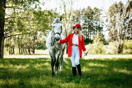 Beautiful young girl with blond hair in a red jacket with a fringe , wearing red hat, smiling and stroking her horse in summer field. equestrian sport conceptの写真素材