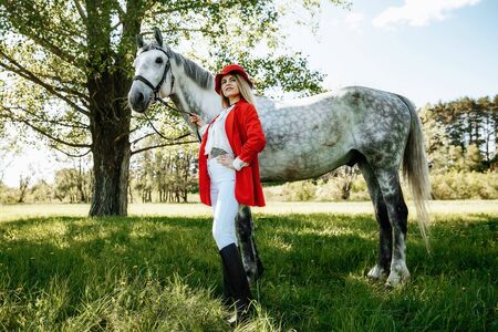 Beautiful young girl with blond hair in a red jacket with a fringe , wearing red hat, smiling and stroking her horse in summer field. equestrian sport conceptの写真素材