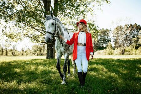 Beautiful young girl with blond hair in a red jacket with a fringe , wearing red hat, smiling and stroking her horse in summer field. equestrian sport conceptの写真素材
